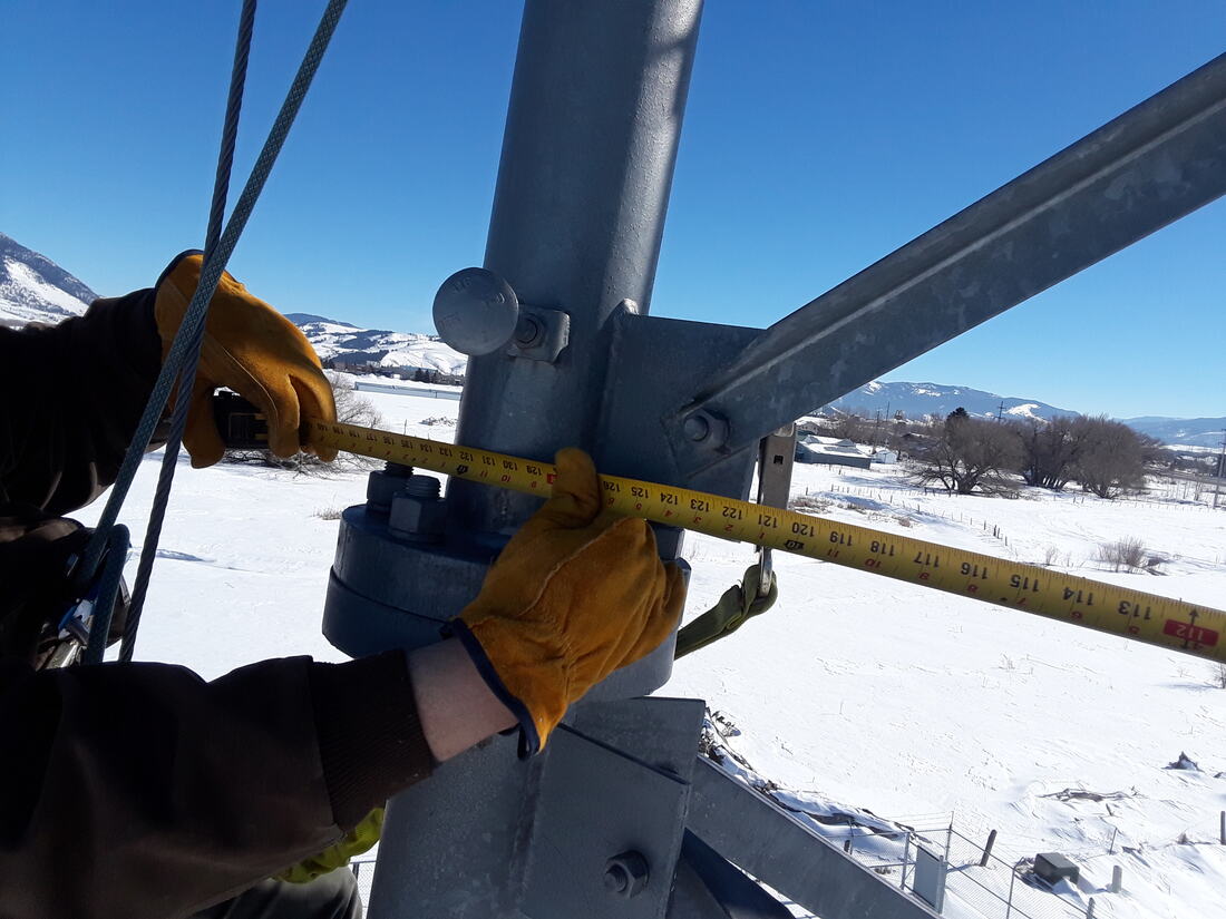 Gloved hands holding a tape measure across a tower member above a snowy valley