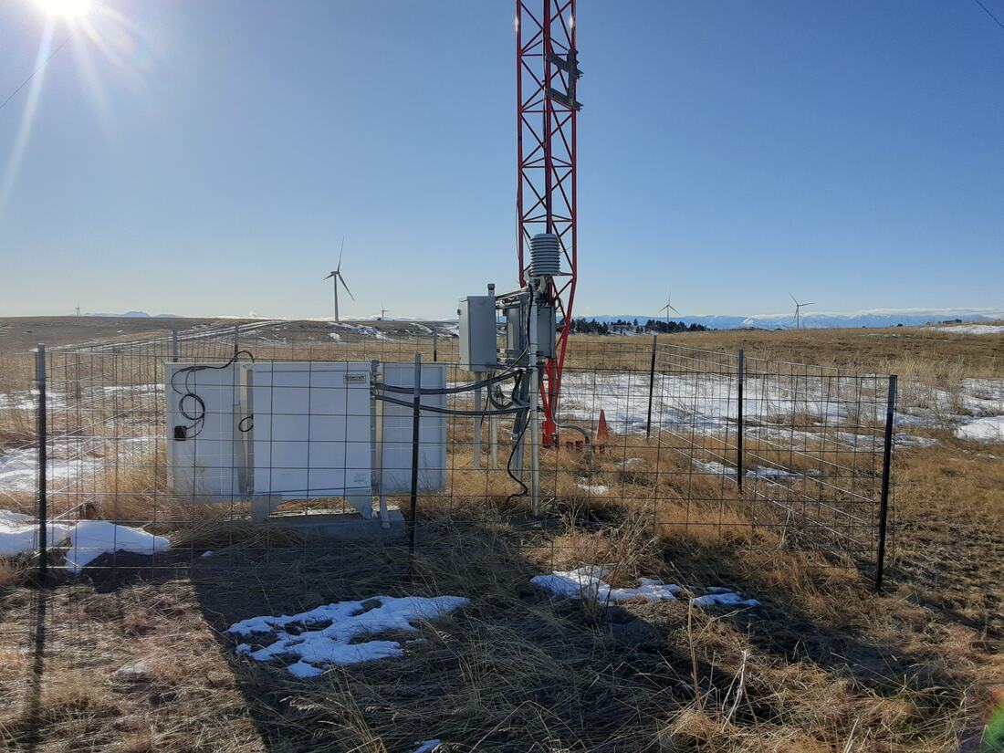 Fenced tower site on the prairie with wind turbines behind